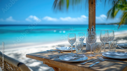 Elegant wooden table set for lunch on white sandy beach, crystal glassware and fine china, turquoise sea in background with clear blue sky, empty space on right 