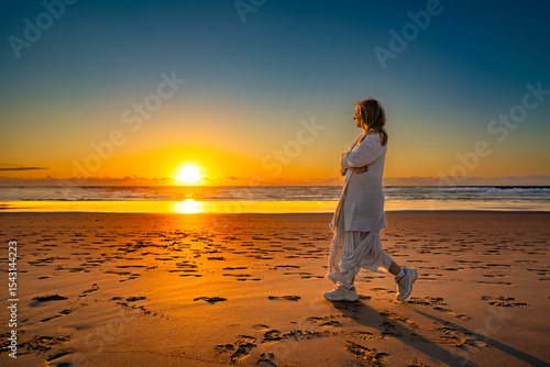Beautiful middle-aged woman walking on sandy beach on sunset on windy spring day. Side view. Monte Clerigo beach on Algarve coast in Portugal