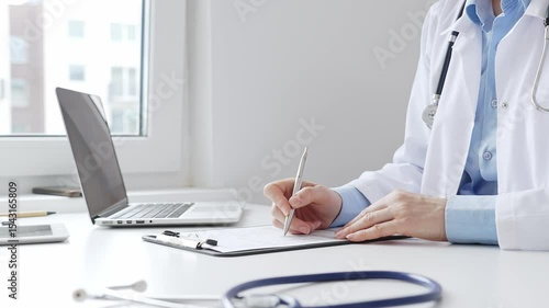 Female doctor completing healthcare paperwork, clipboard positioned near laptop and stethoscope in well lit clinical workspace by window. Medicine and health care concept