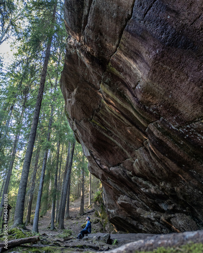 Dramatic rock formation at Paistjärvi’s Pirunkirkko (Devil’s Church), a natural cliff in a Finnish forest. A person sits below the massive boulder, highlighting the scale of the geological feature. 