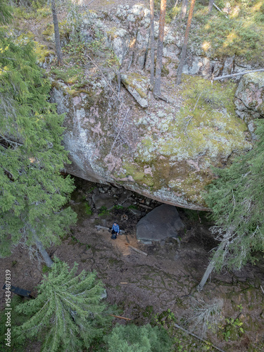 Aerial view of a person standing beneath the dramatic rock overhang of Pirunkirkko (Devil’s Church) in Paistjärvi, Finland. The moss-covered cliffs and surrounding pine forest.
