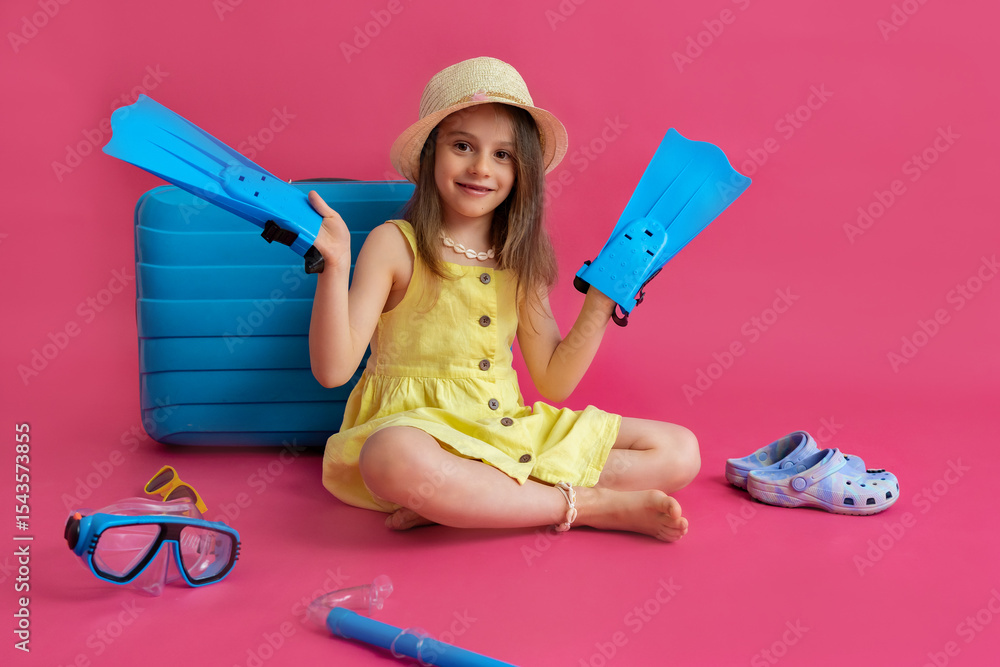 Little girl in a panama hat sitting in front of a blue suitcase and holding blue flippers on a pink background with space for text. Concept of travel, flight, vacation, relaxation. Little girl in a panama hat sitting in front of a blue suitcase and holding blue flippers on a pink background with space for text. Concept of travel, flight, vacation, relaxation.