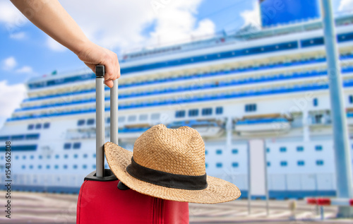 For travel and vacations purposes, a simple modern suitcase in red color at ship in a port with a straw hat.