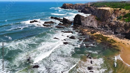 Wide aerial drone shot moving over large rolling waves and a rugged rocky beach with towering cliffs at Praia da Amália, Costa Vicentina, Portugal. Showcasing the powerful ocean meeting dramatic coast