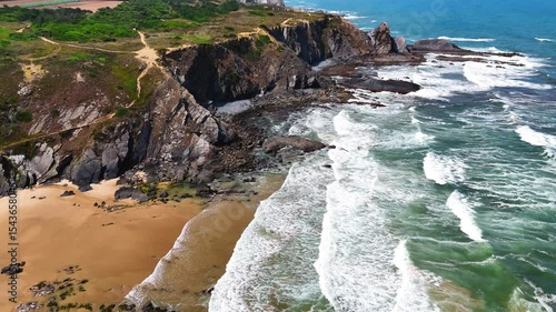 Wide aerial drone shot moving over large rolling waves and a rugged rocky beach with towering cliffs at Praia da Amália, Costa Vicentina, Portugal. Showcasing the powerful ocean meeting dramatic coast