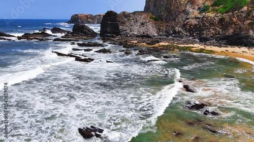 Moving aerial view following ocean waves as they roll and crash along the shore at Praia da Amália, Costa Vicentina, Portugal. Showcasing the energy and flow of the coastal scene.