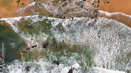 Top-down aerial view of ocean waves crashing onto the sand at Praia da Amália, Costa Vicentina, Portugal. Captured in slow motion, showing a dramatic coastal scene.