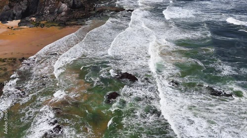 Moving aerial view following ocean waves as they roll and crash along the shore at Praia da Amália, Costa Vicentina, Portugal. Showcasing the energy and flow of the coastal scene.