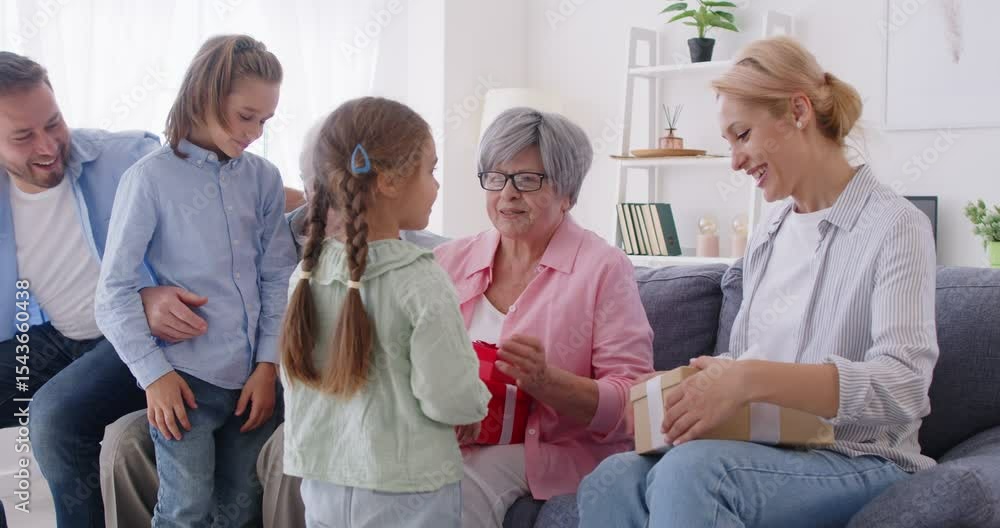 Two lovely children boy and girl congratulating their grandmother with birthday and giving her gift boxes. Happy big multi generation family sitting on sofa, celebrating special occasion together.