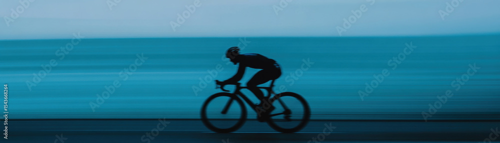 Fototapeta premium Cyclist in aerodynamic skinsuit glides effortlessly along coastal road, surrounded by serene blue backdrop. motion captures essence of speed and tranquility