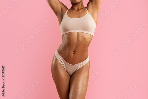 Cropped studio shot of African American woman in nude underwear standing with arms up, showing her midsection and curves, wellness and healthy lifestyle concept.