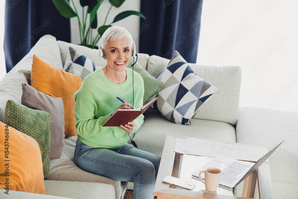 Obraz premium Confident woman with white hair enjoying leisure time at home, taking notes on a sofa with a laptop and a headset