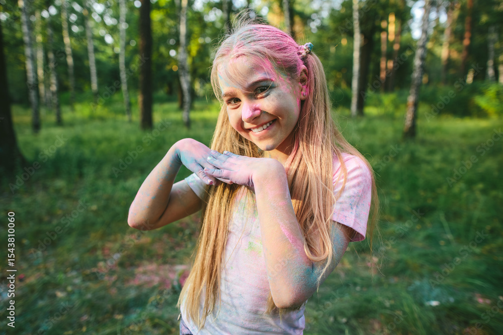 Naklejka premium Caucasian tween girl with long blond hair and colorful powder on face and arms poses playfully in summer forest, expressing joy, spontaneity, and carefree childhood celebration outdoors 