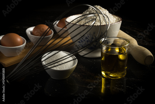 Ingredientes para hornear, incluyendo huevos, harina, azúcar y aceite, dispuestos con utensilios de cocina sobre un fondo oscuro. 