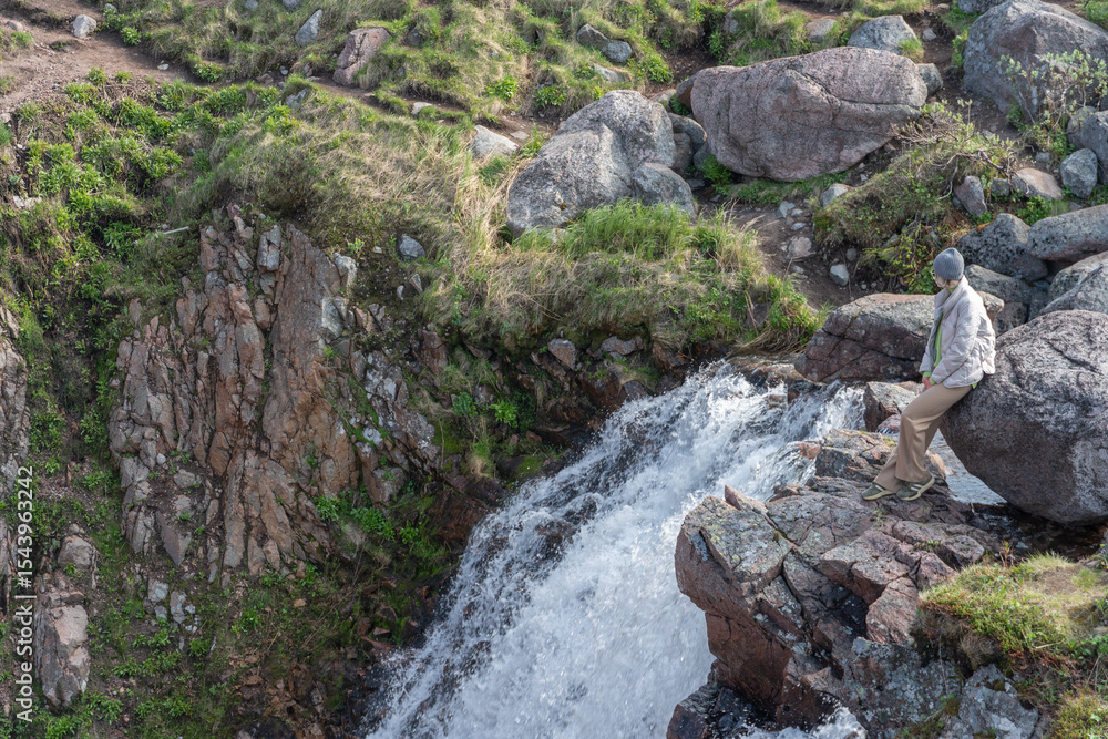 Woman in light winter clothing sits relaxed on a rock next to a flowing waterfall in Teriberka, Russia, surrounded by rugged stone formations Woman in light winter clothing sits relaxed on a rock next to a flowing waterfall in Teriberka, Russia, surrounded by rugged stone formations