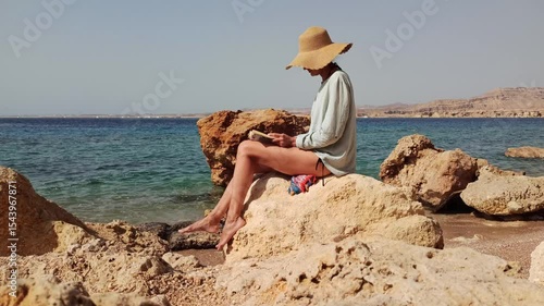 A young woman sits on a stone by the sea and reads a book. She has a straw sunhat. The clear blue water and rocky beach create a tranquil atmosphere.