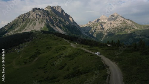 White van driving on winding mountain road surrounded by lush green landscapes and majestic peaks, aerial view