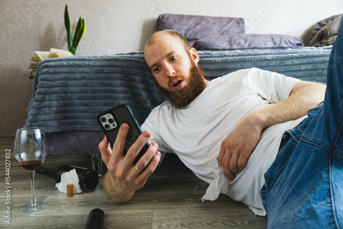 Surprised bearded man lying on the floor, holding smartphone and staring at screen with shocked expression. Messy scene with wine glass, bottle, and tissues suggests emotional or dramatic situation.

