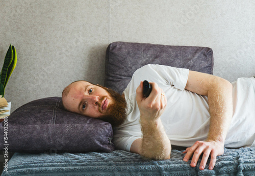 Unmotivated bearded man lying in bed with remote control, looking bored and uninterested. Concept of lazy day, boredom, or passive leisure at home.

