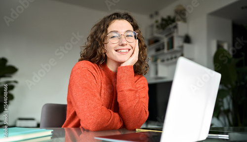 Portrait of happy young woman or student wearing glasses sitting at desk at home office with laptop computer looking at camera advertising online learning, remote work, business webinars.