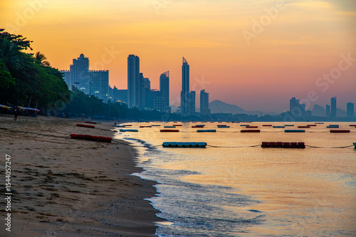 Dawn view of Jomtien Beach, Pattaya, Thailand. City skyline and distant hills silhouetted against a colorful sunrise sky. Morning mist and streetlights still glowing.

