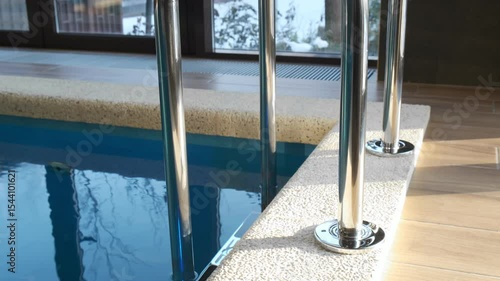 Metal handrails at the edge of an indoor overflow pool with clear blue water. Modern finish and a winter landscape outside the window. Cozy relaxing atmosphere
