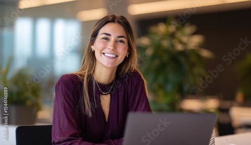 Smiling woman with a laptop in an office setting wearing a purple blouse