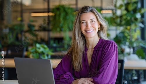 Smiling woman with long hair sits at a desk with a laptop in a plantfilled office