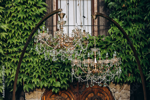Two ornate crystal chandeliers suspended outdoors in front of an ivy-covered wall with a gothic arched window, creating a majestic and romantic scene.