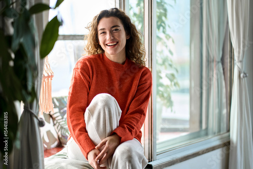 Happy pretty young positive charming woman sitting on windowsill relaxing at home looking at camera feeling joy. Smiling cheerful lady chilling in apartment, laughing, having fun, portrait.