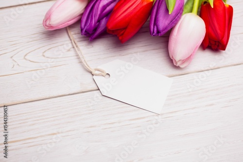 Fototapeta Naklejka Na Ścianę i Meble -  Flat design is showing tulip blossoms resting on pale wooden tabletop with blank white gift tag