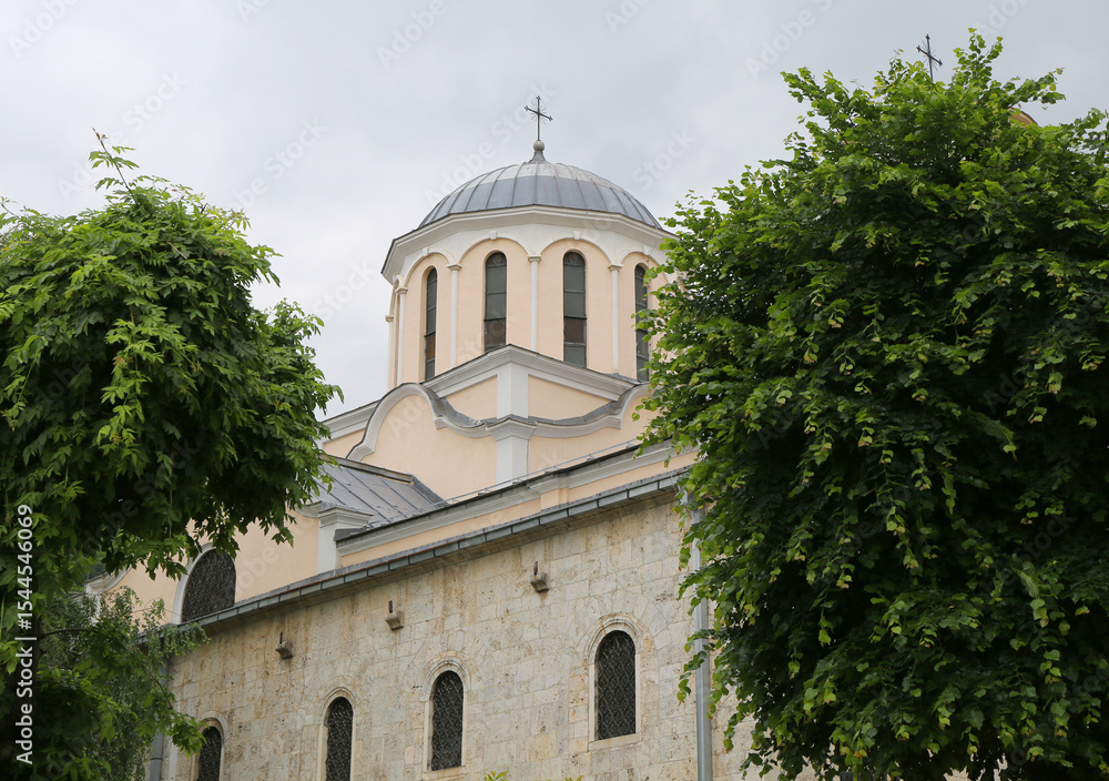Fototapeta premium Dome with cross of St George Cathedral building between tree and plants in Prizren, Kosovo