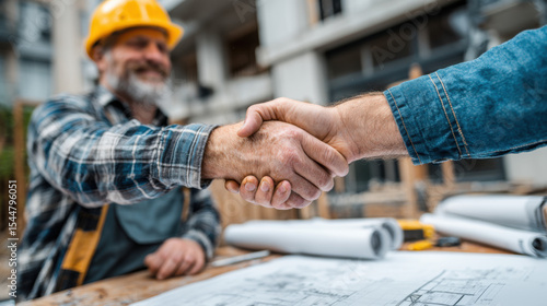 Construction worker wea a hard hat shakes hands with a client over blueprints at a construction site after successful agreement and future project planning.