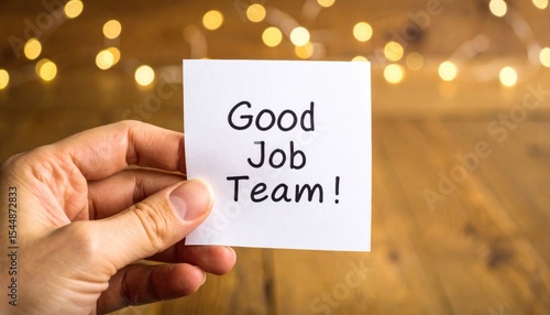 Hand holds sticky note with 'Good Job Team!' message, warm background lights convey celebration