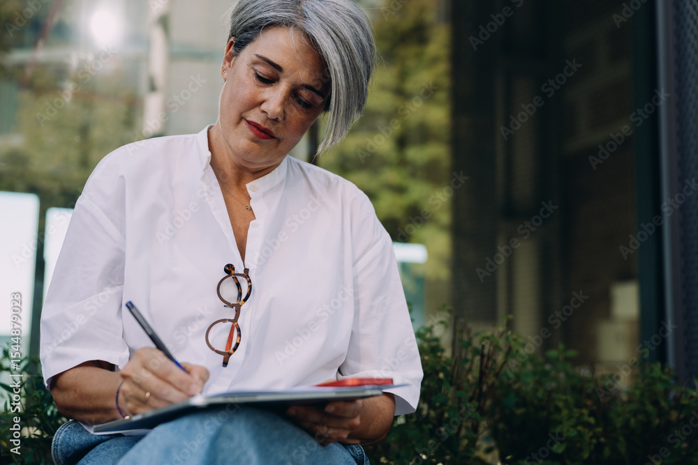 Confident senior woman writing with focus in notebook while sitting outside, working on creative ideas or business planning in modern city environment, enjoying calm and productivity Confident senior woman writing with focus in notebook while sitting outside, working on creative ideas or business planning in modern city environment, enjoying calm and productivity