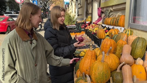 Two women are examining pumpkins at an outdoor market stall.  Autumn harvest abundance of various gourds, apples and grapes are visible. A vibrant autumn scene.