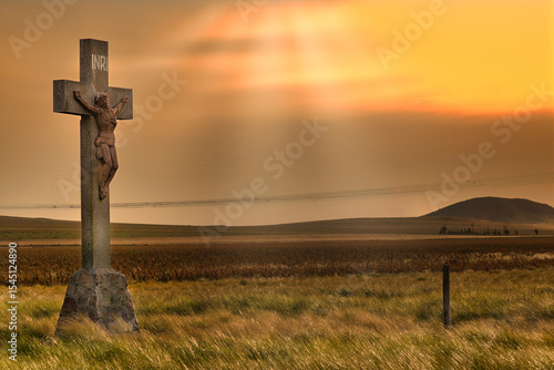 Fototapeta Large crucifix stands in field at sunset with power lines
