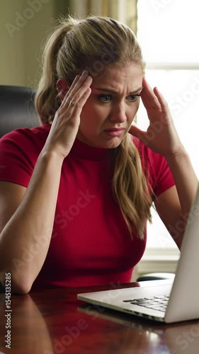Young caucasian woman, mid-20s, in red top, hunched at dark wood desk in sunlit home office, deeply stressed by laptop's plummeting financial graph, slowly presses hands to temples, overwhelmed with