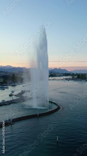 Aerial wide video of Geneva's Jet d'Eau fountain powerfully spraying water at twilight, catching golden hour glow. Drone orbits Lake Geneva's deep turquoise water, revealing twinkling city skyline