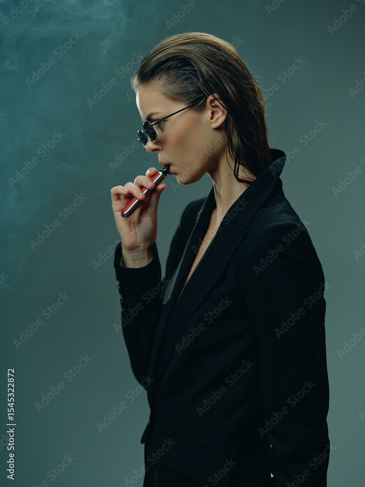 Obraz premium Woman vaping holds electronic cigarette with smoke around, wearing black suit and sunglasses in moody atmosphere, indoor setting with dark background and stylish look.