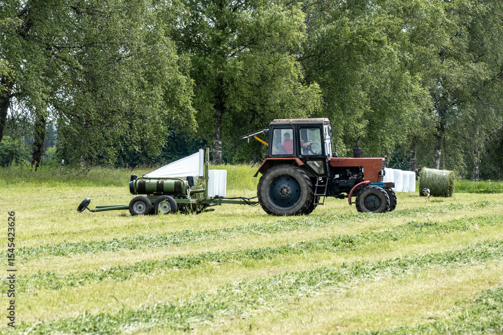 Fototapeta premium Tractor Wrapping Hay Bales with White Film in Green Meadow – Modern Agriculture in Action