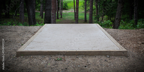 Close-up of a concrete tee pad on a disc golf course with a basket in the background. A stable and level platform made of concrete. pine tree landscape.