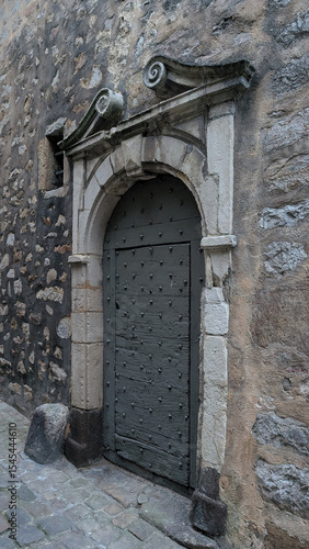 Ancient medieval wooden door with metal studs framed by a stone arch in a wall of stone masonry off a cobblestone road