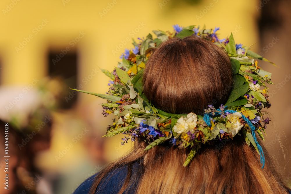 Naklejka premium Close-up of a flower crown made of colorful wildflowers and greenery on a woman's brown hair, viewed from behind during a traditional outdoor celebration.
