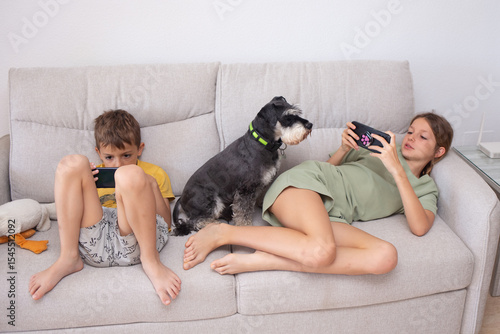 Children enjoy mobile games while a bored dog sits beside them on a couch