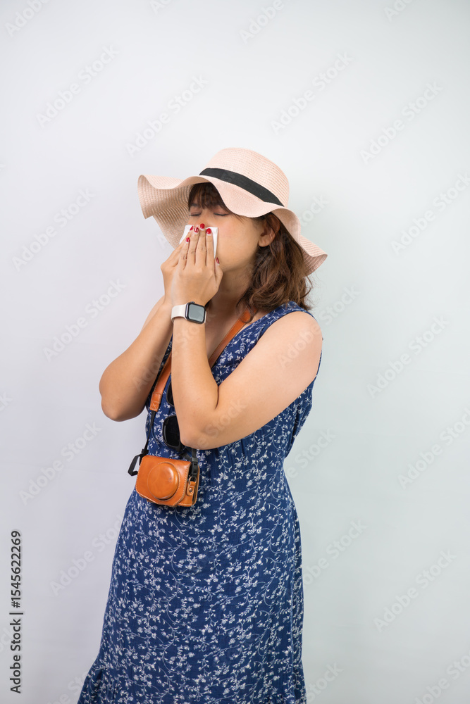 Fototapeta premium Beautiful young asian woman wearing blue summer dress and beach straw hat is sneezing and covering using tissue paper, isolated over white background.