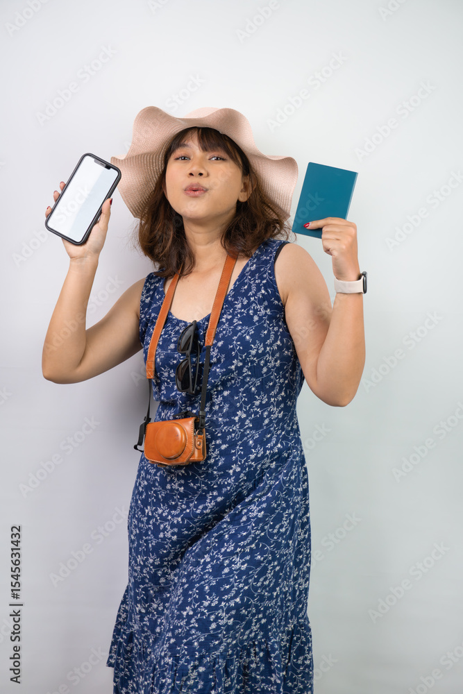 Naklejka premium Beautiful young asian woman wearing blue summer dress and beach straw hat is holding smartphone with blank screen and indonesian passport, isolated over white background.