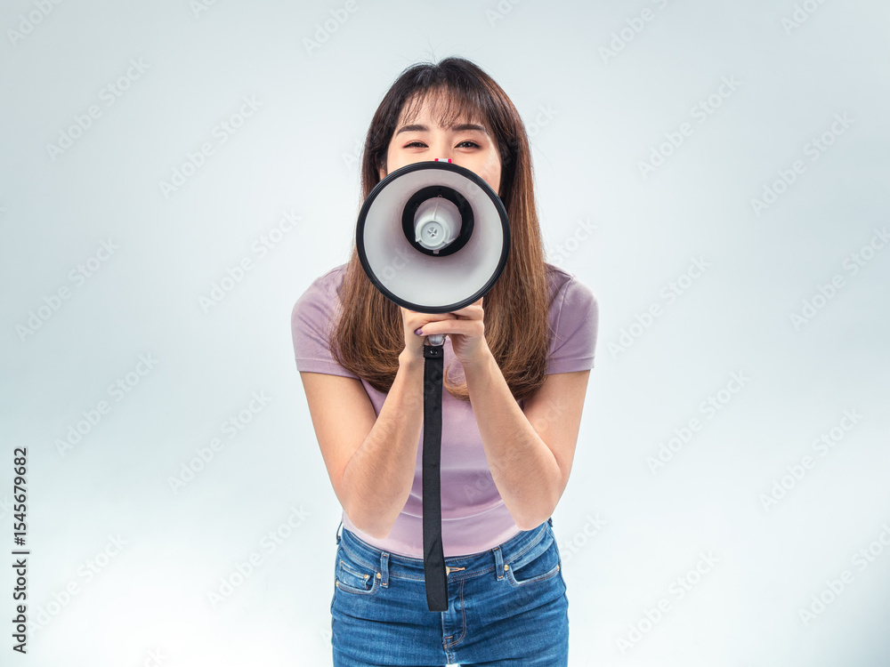Fototapeta premium Asian woman in purple t-shirt and jeans shouting into a white and red megaphone, expressing urgency or announcement while standing in studio with plain white background