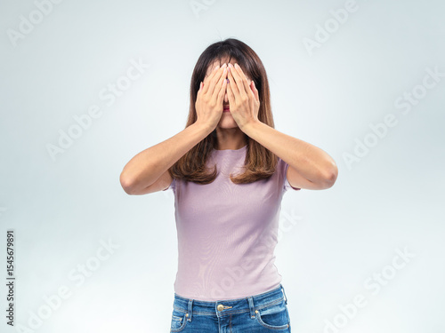 Asian woman in purple t-shirt and jeans covering her face with both hands, standing in studio against plain white background, showing expression of shyness, fear, or embarrassment
