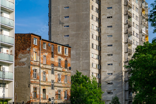 Architectural contrast in Łódź – a dilapidated brick tenement with peeling plaster surrounded by modern concrete and glass apartment buildings. Greenery in the foreground.